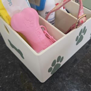 May include: A white plastic storage caddy with multiple compartments, decorated with green paw print decals. Inside are various pet care items, including a pink bone-shaped waste bag dispenser with the name Bella printed on the side.