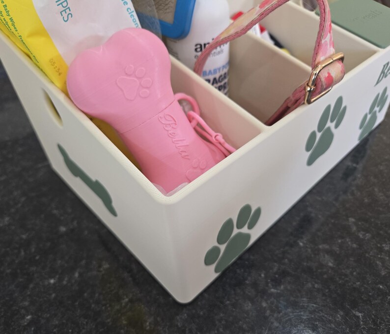 May include: A white plastic storage caddy with multiple compartments, decorated with green paw print decals. Inside are various pet care items, including a pink bone-shaped waste bag dispenser with the name Bella printed on the side.