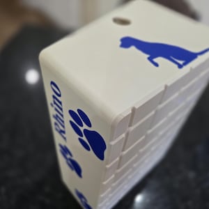 May include: A rectangular off-white box featuring a blue silhouette of a dog on the top surface. The side panel displays the name Rhino in blue text, accompanied by blue paw print graphics. The box has a grid-like textured pattern on the side.