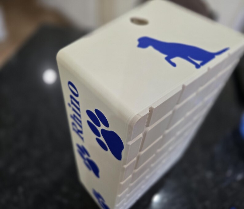 May include: A rectangular off-white box featuring a blue silhouette of a dog on the top surface. The side panel displays the name Rhino in blue text, accompanied by blue paw print graphics. The box has a grid-like textured pattern on the side.