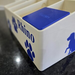 May include: A white rectangular storage organizer with multiple compartments and a blue lid. The side features blue graphics of a paw print and a dog silhouette, along with the text Rhino printed in a bold blue font on the front panel.