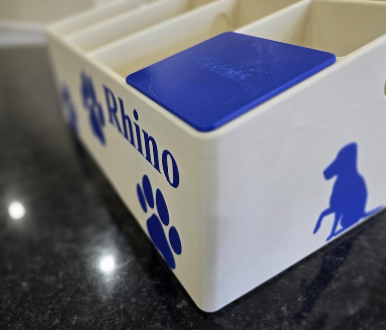 May include: A white rectangular storage organizer with multiple compartments and a blue lid. The side features blue graphics of a paw print and a dog silhouette, along with the text Rhino printed in a bold blue font on the front panel.