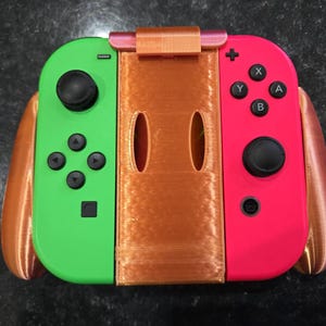 May include: A green and red game controller with black buttons and a brown center piece. The controller has a brown bracket on top and brown extensions on the sides.