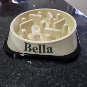 May include: A cream-colored slow feeder pet bowl with a black base. The interior features a series of raised ridges and obstacles to slow down eating. The name Bella is printed in dark green lettering on the front side of the bowl.