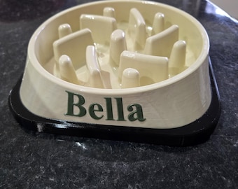May include: A cream-colored slow feeder pet bowl with a black base. The interior features a series of raised ridges and obstacles to slow down eating. The name Bella is printed in dark green lettering on the front side of the bowl.