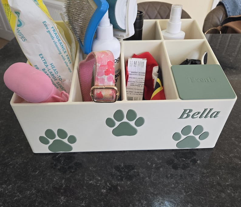 May include: A cream-colored rectangular pet supply organizer with multiple compartments. The front features three sage green paw print decals and the name Bella in script. A sage green lid labeled Treats covers one section, holding various pet care items.