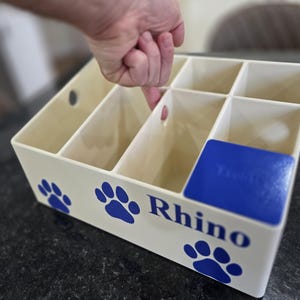 May include: A cream-colored plastic storage organizer with multiple compartments and circular cutouts. The front features blue paw print graphics and the word Rhino printed in blue. A blue square insert is positioned in one corner compartment.