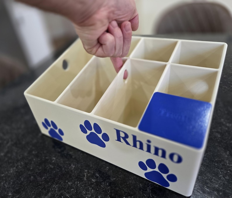 May include: A cream-colored plastic storage organizer with multiple compartments and circular cutouts. The front features blue paw print graphics and the word Rhino printed in blue. A blue square insert is positioned in one corner compartment.