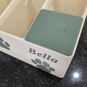 May include: A white plastic storage container with multiple compartments, featuring sage green paw print accents and the name Bella printed on the front. A matching sage green lid with the word Treats embossed on it covers one of the sections.