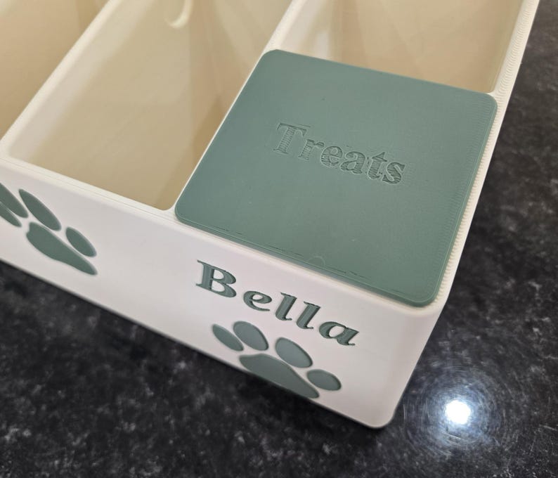 May include: A white plastic storage container with multiple compartments, featuring sage green paw print accents and the name Bella printed on the front. A matching sage green lid with the word Treats embossed on it covers one of the sections.