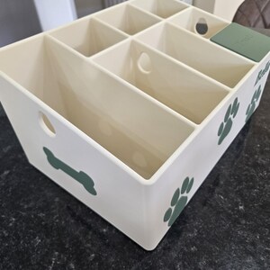 May include: A cream-colored plastic storage organizer with multiple compartments, featuring dark green decals of paw prints, a bone, and the name Bella. A small dark green lid labeled Treats covers one section, and circular cutouts appear on the sides.
