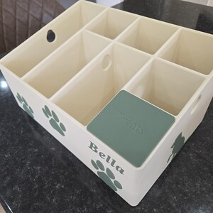 May include: A cream-colored plastic storage organizer with six compartments, featuring green paw print decals on the exterior. The front side displays the name Bella in green lettering, and one compartment is covered by a green lid labeled Treats.