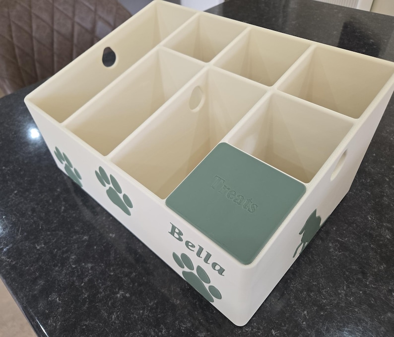 May include: A cream-colored plastic storage organizer with six compartments, featuring green paw print decals on the exterior. The front side displays the name Bella in green lettering, and one compartment is covered by a green lid labeled Treats.