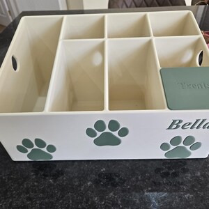 May include: A cream-colored plastic storage organizer with multiple compartments, featuring green paw print decals on the front. The name Bella is printed in green on the front, and a green lid labeled Treats covers one of the smaller compartments.