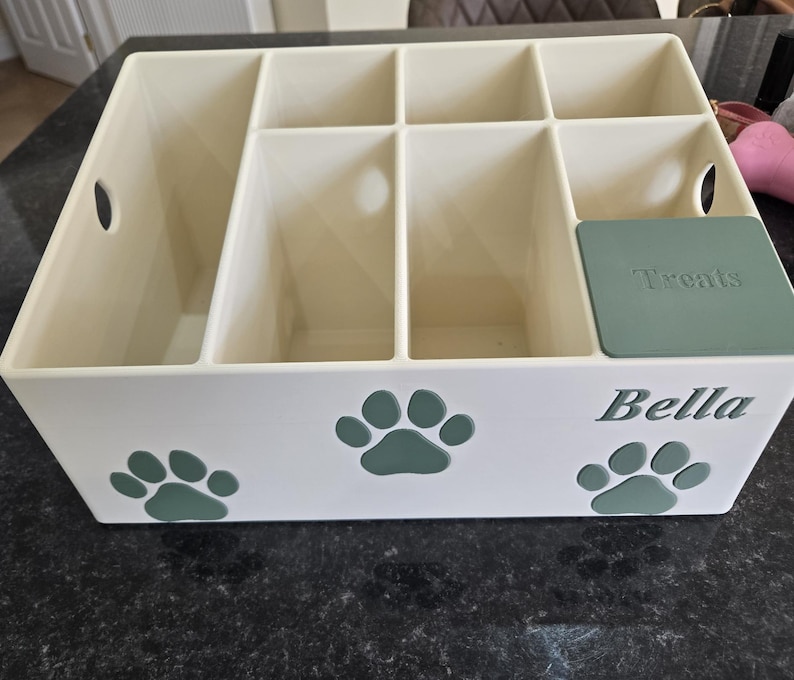 May include: A cream-colored plastic storage organizer with multiple compartments, featuring green paw print decals on the front. The name Bella is printed in green on the front, and a green lid labeled Treats covers one of the smaller compartments.