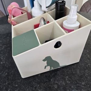 May include: A cream-colored plastic storage caddy with multiple compartments, featuring a green silhouette of a dog on the front and a green lid labeled "Treats" covering one section. The caddy contains various items including bottles and a pink strap.