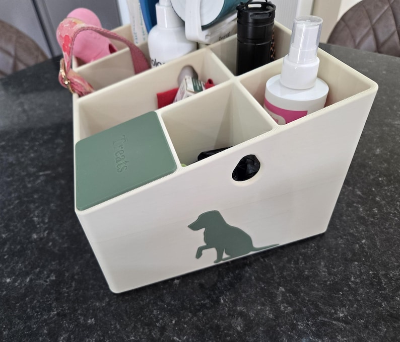 May include: A cream-colored plastic storage caddy with multiple compartments, featuring a green silhouette of a dog on the front and a green lid labeled "Treats" covering one section. The caddy contains various items including bottles and a pink strap.