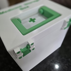 May include: A white medicine storage box featuring a light green handle and a matching green latch mechanism. The top surface displays a green cross symbol and the text Medicine Storage Box printed in green. The box has a square, boxy shape.