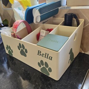 May include: A cream-colored plastic storage organizer with multiple compartments, featuring green paw print decals, a silhouette of a dog, and the name Bella printed on the front. A green lid labeled Treats covers one section.