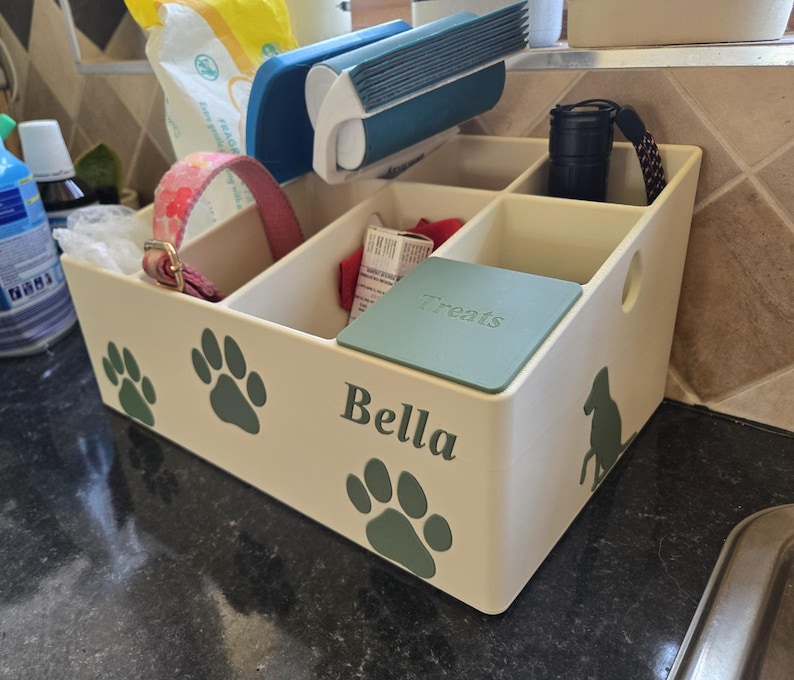 May include: A cream-colored plastic storage organizer with multiple compartments, featuring green paw print decals, a silhouette of a dog, and the name Bella printed on the front. A green lid labeled Treats covers one section.