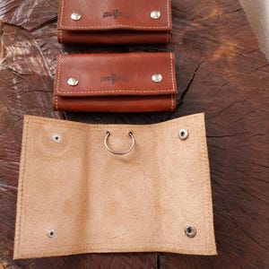 May include: Three brown leather key holders on a wooden surface. Two are closed, showing the "JOW WOLF" logo and silver snap closures. The third is open, revealing a lighter interior and a metal ring. The key holders are stitched with a contrasting thread.