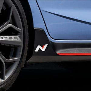 May include: Close-up of a car's side panel featuring a black accent with a white "N" logo and a red stripe. The car's blue body and a portion of the wheel are visible against a dark background.