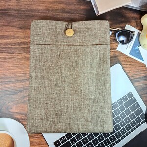 以下が含まれることがあります： ボタン留めの茶色のリネン製ラップトップスリーブ。スリーブは木製の表面に置かれ、背景にはラップトップのキーボードが見えます。