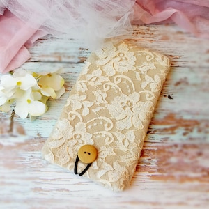 Puede incluir: Una funda para teléfono de tela beige con un patrón de encaje blanco y un cierre de botón de madera.