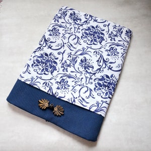 Puede incluir: Una cubierta de cuaderno con estampado azul y blanco, con una solapa azul marino y dos detalles decorativos de metal dorado.