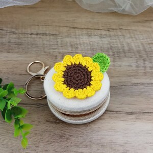 May include: A small white zippered pouch with a crocheted sunflower design. The sunflower is yellow with a brown centre and a green leaf. The pouch has a gold metal key ring attached.
