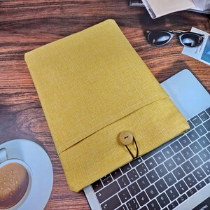 以下が含まれることがあります： 木製のボタン留めが付いた黄色のリネン製のラップトップスリーブ。スリーブはラップトップのキーボードの上に置かれています。