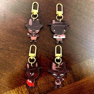 Op de afbeelding: Vier sleutelhangers met goudkleurige sluitingen. Elke sleutelhanger heeft een cartoonfiguur met een hoed en verschillende outfits. De figuren zijn voornamelijk bruin met rode, witte en zwarte accenten. De sleutelhangers worden op een houten oppervlak weergegeven.
