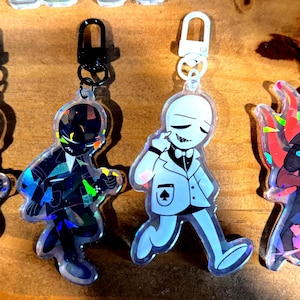 Puede incluir: Cuatro llaveros acrílicos con figuras de dibujos animados con trajes. Los llaveros tienen diseños negros, blancos e iridiscentes. Cada llavero tiene un cierre de diferente color: negro, negro, blanco y rojo. Las figuras tienen un tema de naipes.