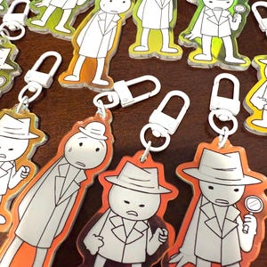 以下が含まれることがあります： 探偵をテーマにしたキーホルダーのコレクション。各キーホルダーには、白いコートと帽子をかぶった漫画の探偵キャラクターが描かれており、オレンジ、黄色、または緑色のボーダーが付いています。虫眼鏡を持っているものもあります。キーホルダーには白い金属製の留め金が付いています。