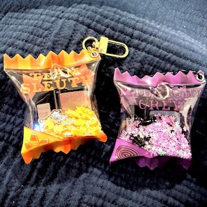 以下が含まれることがあります： オレンジと紫の、カラフルな紙吹雪が入った透明なビニール袋のチャームが2つ。オレンジ色のバッグには「Team Sleuth」、紫色のバッグには「Midnight Crew」と書かれています。どちらも金色のキーホルダーが付いています。