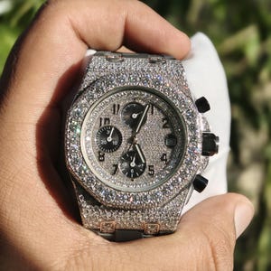 May include: A luxury wristwatch with a silver face and band, adorned with many small, sparkling gemstones. The watch has three sub-dials and black accents on the side buttons. The watch is held in a hand against a blurred green backdrop.