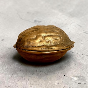 Könnte beinhalten: Eine dekorative Box aus goldfarbenem Metall in Form einer Walnuss. Das Objekt weist eine texturierte, unebene Oberfläche auf, die die natürlichen Rillen und Vertiefungen einer echten Walnussschale nachahmt, mit einer horizontalen Naht.