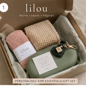 Op de afbeelding: Cadeaudoos met een opgerolde roze handdoek, een beige geweven vierkant, een groene etui met een gouden sleutelhanger en een kleine kaart. De doos heeft het woord "lilou" en de zin "move reset repeat" erop gedrukt. De geschenkset is gelabeld "PERSONALISED GYM ESSENTIALS GIFT SET".