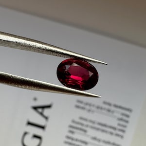 May include: An oval-cut, deep red gemstone held by metal tweezers. Behind the gemstone is a white GIA report document with black text and the GIA logo visible. The gemstone exhibits a faceted surface that reflects light, showing a rich, saturated color.