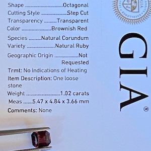 May include: A brownish-red, octagonal-shaped natural ruby gemstone, held by tweezers, is displayed against a GIA report. The report details the stone's weight of 1.02 carats and dimensions of 5.47 x 4.84 x 3.66 mm.