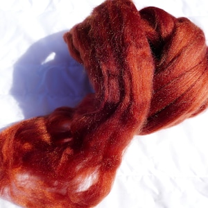 May include: A mass of vibrant, reddish-brown wool roving, ideal for fiber art projects. The wool is soft and fluffy, with a rich, warm color. The fibers are loosely spun, perfect for felting, spinning, or other textile crafts.