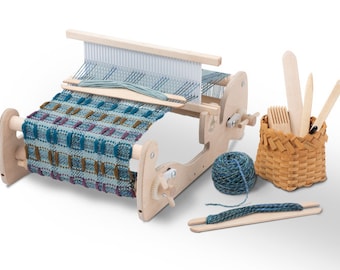 May include: A wooden loom with a partially woven blue, green, and brown tapestry. The loom has a crank and a shuttle. There is a ball of blue yarn, a wooden needle, and a small basket with weaving tools.