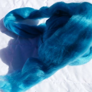 May include: A bright turquoise blue fiber roving, a type of wool used for spinning and felting, is shown in a loose pile on a white surface.