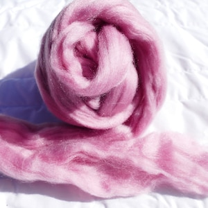 May include: A ball of pink wool roving, a type of fiber used for felting and spinning, is shown on a white surface. The roving is soft and fluffy, and it has a slight sheen.