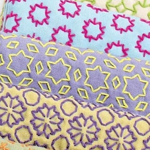 May include: Three rectangular fabric pieces featuring embroidery. The top piece is light blue with pink star-like shapes. The middle piece is lavender with yellow star and diamond patterns. The bottom piece is pale yellow with purple floral motifs.