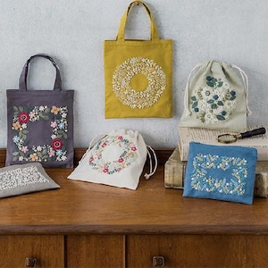May include: A collection of textile items including two tote bags in mustard yellow and dark gray, two drawstring pouches in cream and light green, and two rectangular zippered pouches in gray and blue, all featuring intricate floral embroidery.