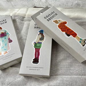Op de afbeelding: Drie rechthoekige lichtgrijze dozen met het label Odekake Brooch Beads Embroidery Brooch Kit. Elke doos bevat een andere kralenillustratie van een persoon in kleurrijke kleding, waaronder een blauwe kimono met patroon, een groene rok en een oranje jas.