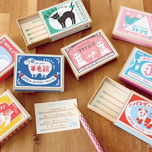 May include: A collection of decorative matchboxes with various designs, including a black cat, sheep, rabbits, and a panda. The boxes are in different colors, such as red, blue, pink, and yellow, with Japanese text and illustrations. Some boxes are open, revealing the matches.