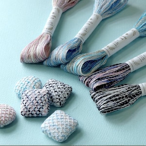 May include: Five skeins of Sashiko lamé embroidery thread with metallic accents, each wrapped in a white paper band, alongside four small, square, and round fabric-covered buttons featuring a cross-stitched grid pattern in various colours.