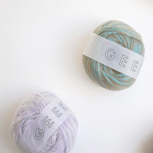 May include: Two balls of yarn with paper labels. One ball is a mix of tan and bright blue fibres with a label reading GEE. The second ball is a soft lavender colour with a label reading GEEK. Both are round, textured, and made of soft material.